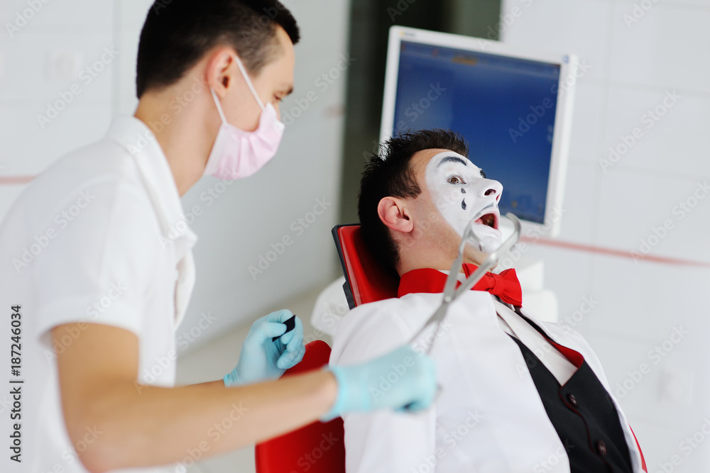 Fototapeta premium The clown or mime is sitting in the dental chair. The dentist treats the tooth to the patient.
