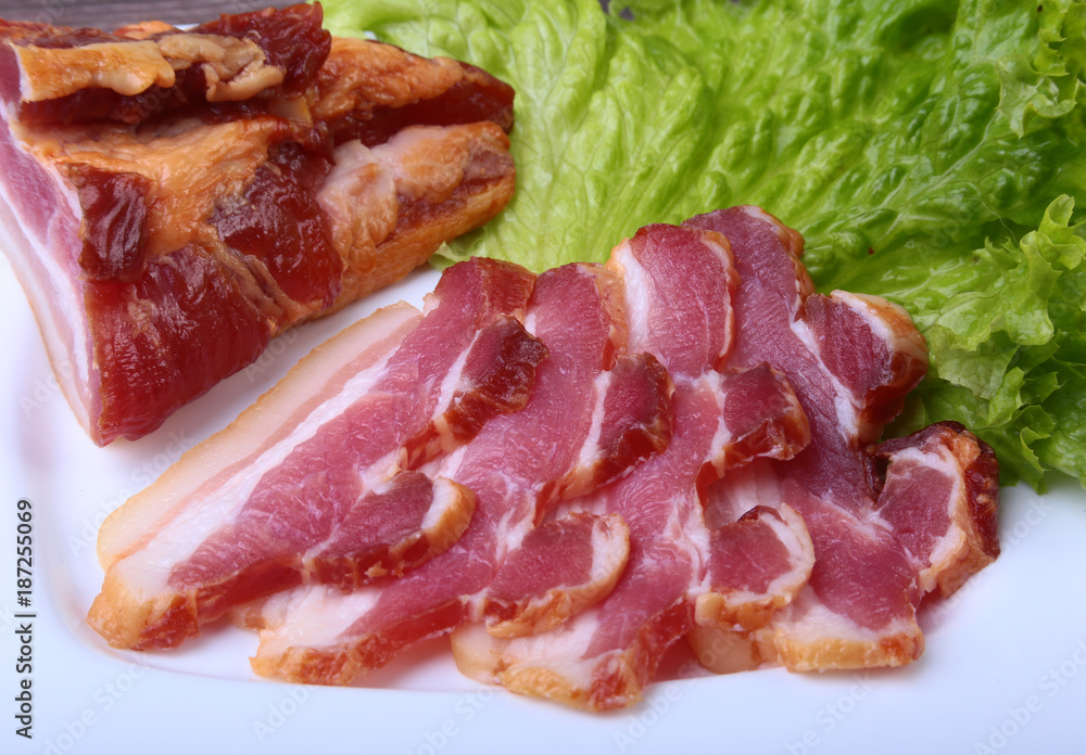 Fresh homemade smoked bacon with leaves lettuce on white plate. Selective focus. Fresh homemade smoked bacon with leaves lettuce on white plate. Selective focus.