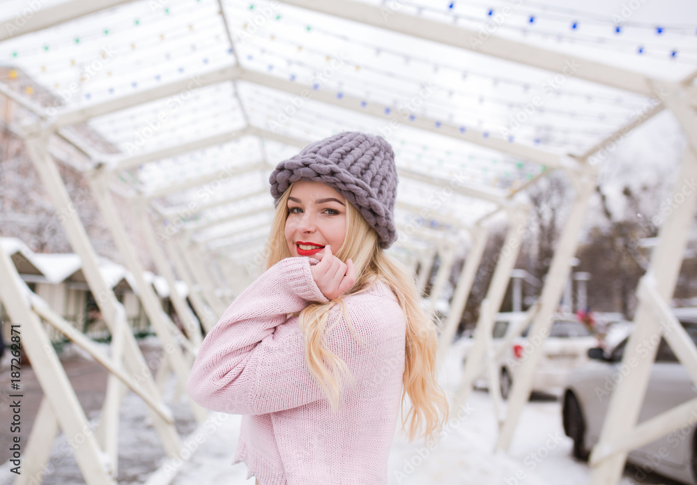 Outdoor portrait of cheerful blonde woman with bright makeup, wearing trendy knitted hat and sweater, posing at the city in winter