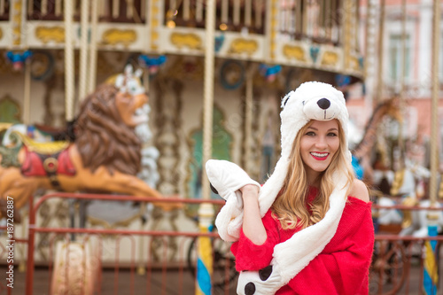 Smiling blonde woman wearing red knitted sweater and funny hat, posing at the background of carousel with lights