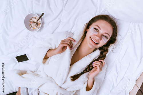 Photography happy young girl in a white terry dressing gown is lying in bed, next to a plate