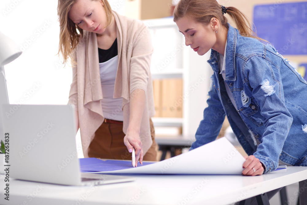 Obraz premium Two young woman standing near desk with instruments, plan and laptop.