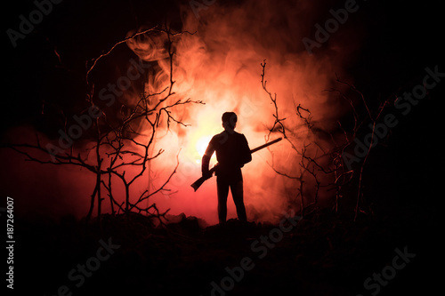 Man with riffle at spooky forest at night with light, or War Concept. Military silhouettes fighting scene on war fog sky background, World War Soldier Silhouette Below Cloudy Skyline At night.