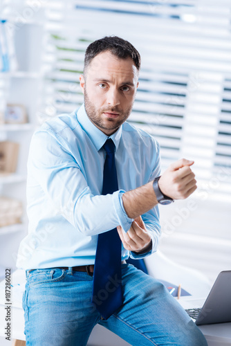 Touching sleeves. Young bearded worker sitting on the table in a comfortable modern office and touching the sleeves of a shirt while preparing to go home after a tiresome day