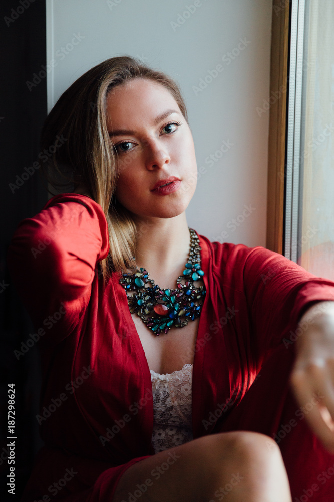beautiful sexy girl in a red robe is sitting on the windowsill at home.  Stock Photo | Adobe Stock