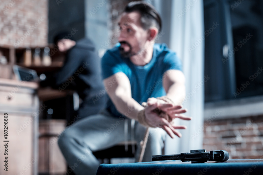 Escaping. Frightened panic bearded man sitting with his hands tied and trying to take the gun while the criminal standing behind him Escaping. Frightened panic bearded man sitting with his hands tied and trying to take the gun while the criminal standing behind him