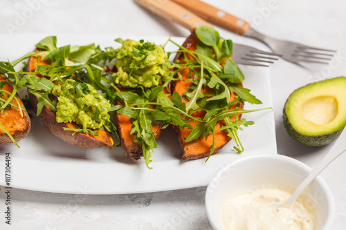 Baked halves of sweet potato with arugula and guacamole. White plate, light background, top view. Healthy vegan food concept.