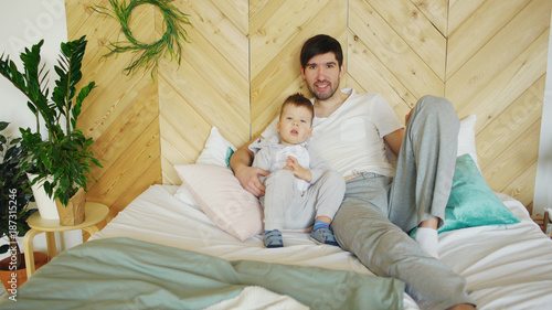 Portrait of happy handsome young father and his son lying on bed smiling and posing into camera