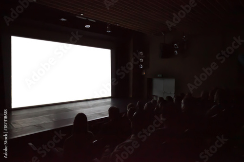 Blurry background of people watching movie in the movie theater with white screen.