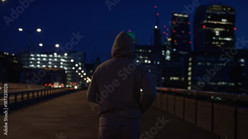 Wide angle shot of camera following a hooded male jogging towards city lights, in slow motion