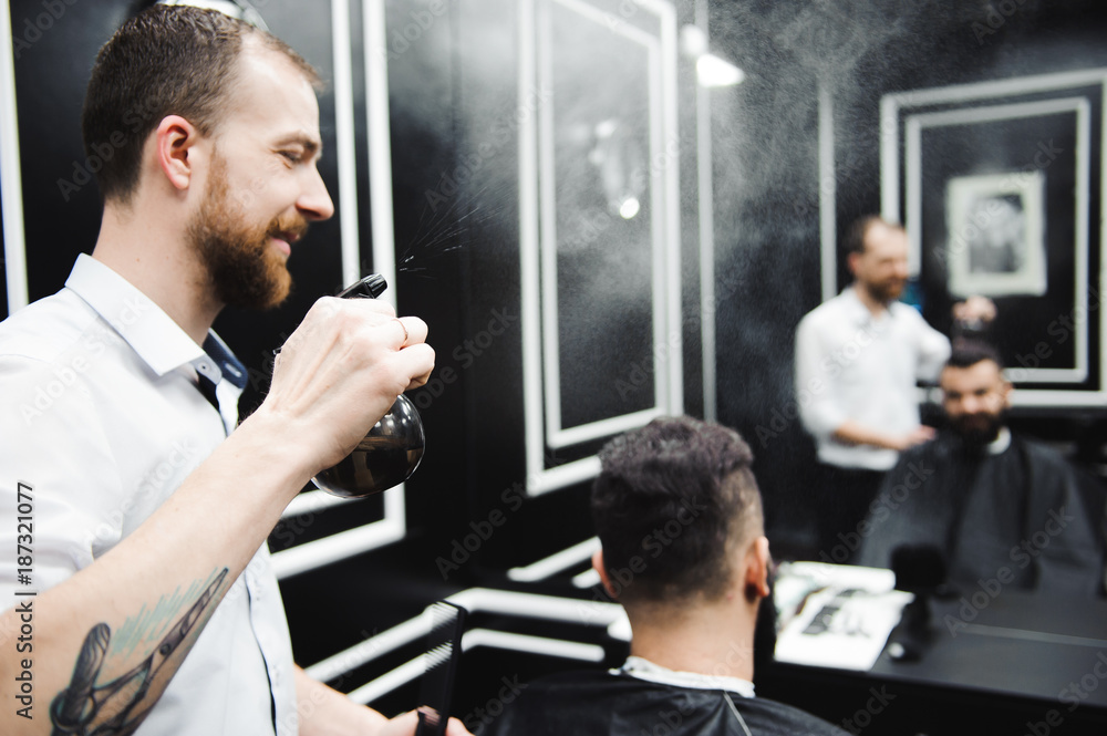 Fototapeta premium Young handsome barber making haircut of attractive man in barbershop