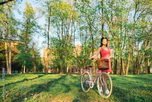 Fashion concept. Lady in a pink dress holding the bike and having nice emotion. A beautiful young woman riding a bicycle with flowers in a basket and enjoying springtime.