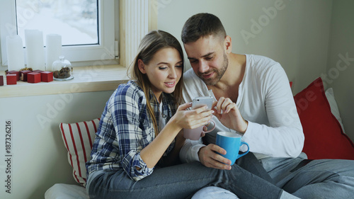 Happy young couple talking and browsing social media on smartphone while sitting in on bed and drink coffe in the morning at home