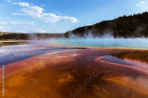 Grand Prismatic Hot Spring In Yellowstone National Park. The deep colors of the spring show the hot temperature. There is evaporating water leaving the spring. It is a sunny day with clouds. 