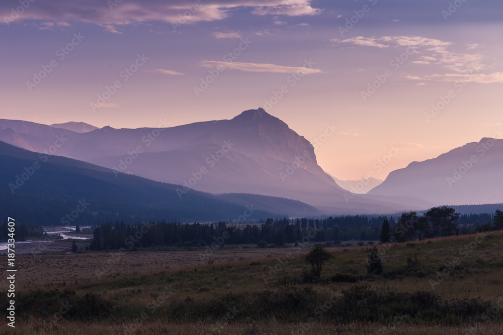 A meadow with a mountain in the background at sunset. There is a river running with a grassy meadow and a haze in the distance. The purple sky shows the different layers of mountains.