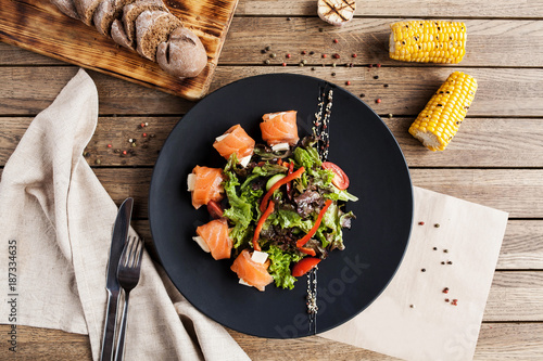 salad with vegetables, cheese and fresh salmon rolls on a black plate with a bread board and corn on a wooden table