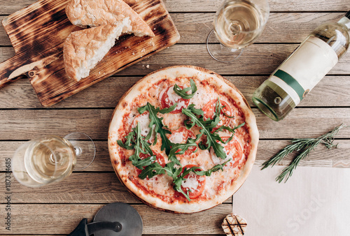 Italian pizza margherita with arugula, with a bottle and two glasses of white wine on a wooden table