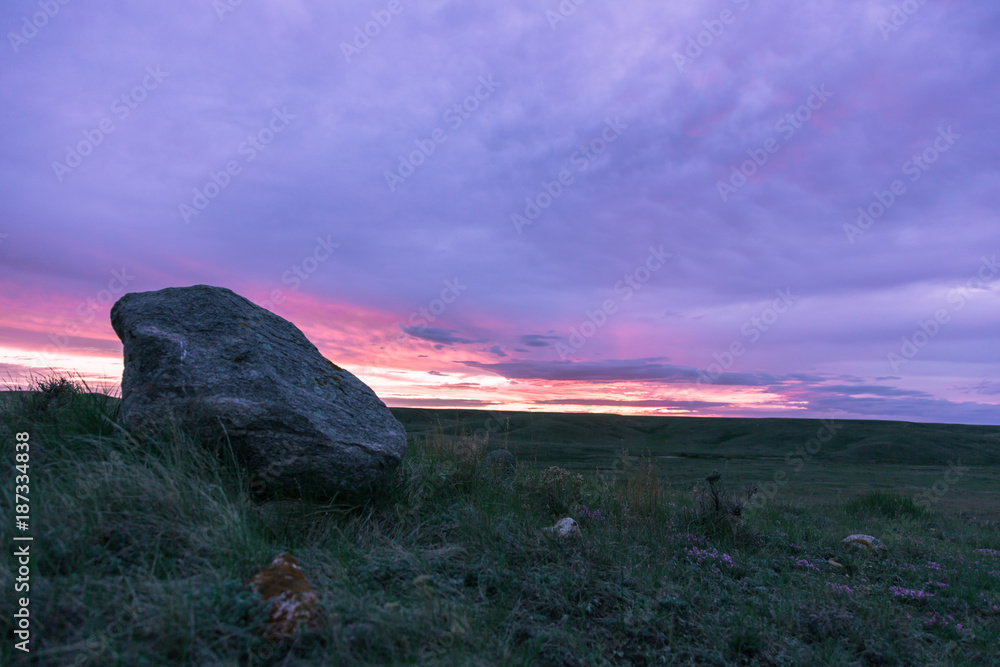Rock at sunset in Grasslands National Park, Saskatchewan. The purple red and orange sky roar across the flat plains of SK. There is a large rock with grass in the foreground.