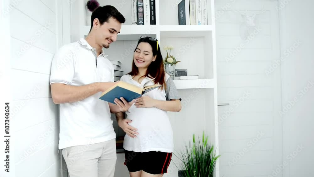 A pregnant asian woman and her white husband lying at home reading a book together in hall way. Discussing, smiling, talking to each other.. Family concept. A pregnant asian woman and her white husband lying at home reading a book together in hall way. Discussing, smiling, talking to each other.. Family concept.