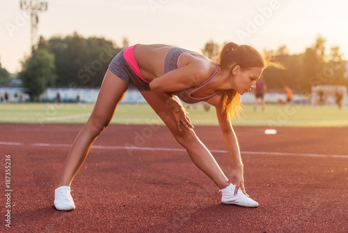 Side view of athletic woman working out in stadium, bending and stretching her back leg muscles