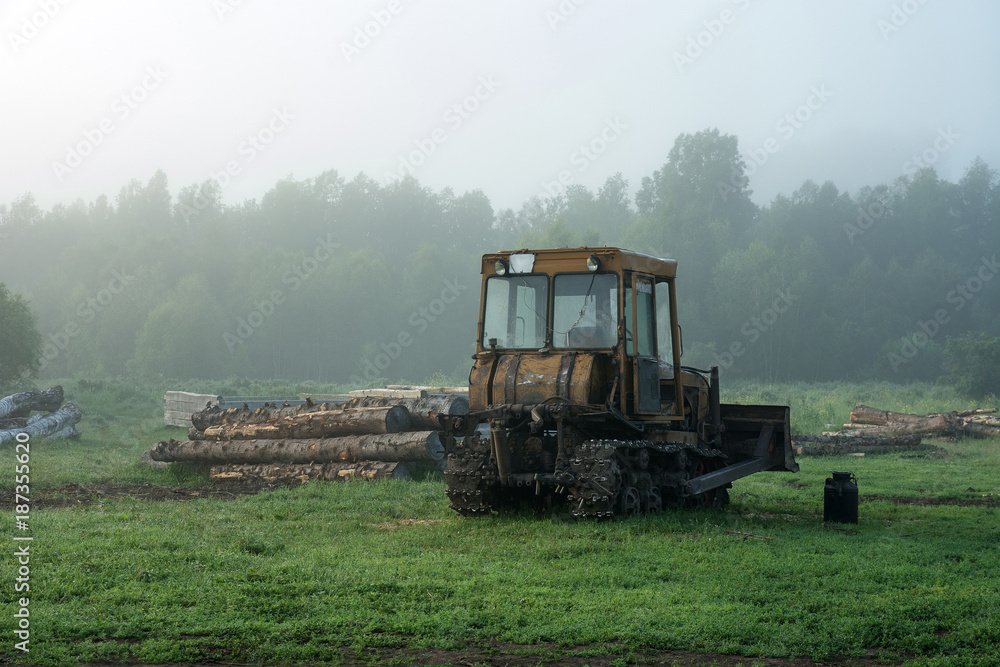 Obraz premium The caterpillar tractor stands in the morning fog on the background of logs and trees. Green grass.