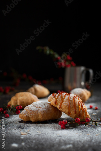 several whole croissants with crumbs, powdered sugar, caramel, a glass with berries and red berries on a dark background