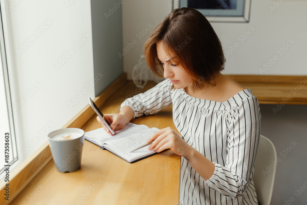 Young freelance girl worker sitting at cafe shop with phone, note book, and thinking about work. Freelance work place. Young freelance girl worker sitting at cafe shop with phone, note book, and thinking about work. Freelance work place.