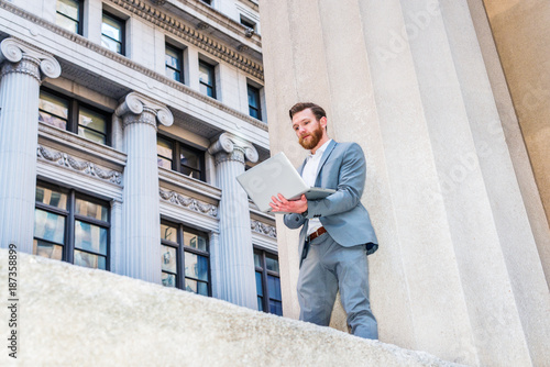 American Businessman with beard, mustache working outside in New York, wearing cadet blue suit, white undershirt, standing against column on street with vintage buildings, working on laptop computer