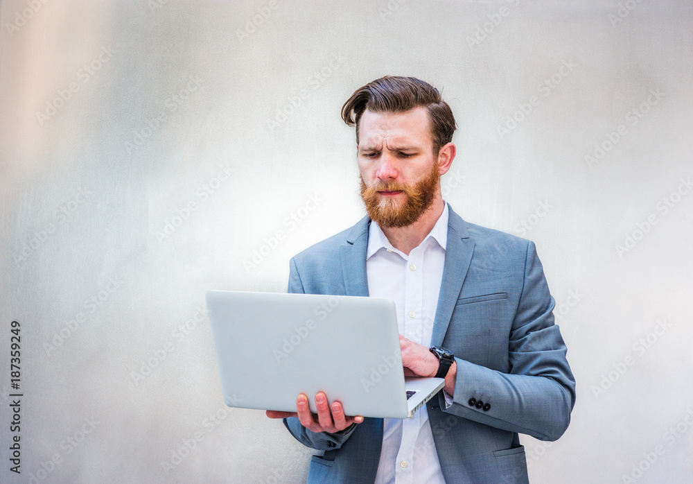 Young American Hipster Businessman with full beard, working on laptop computer, on silver metal background.. Young American Hipster Businessman with full beard, working on laptop computer, on silver metal background..