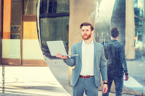 Εκτύπωση καμβά Young American Hipster Businessman with full beard working in New York, wearing cadet blue suit, white shirt, standing by metal mirror wall, holding laptop computer, thinking