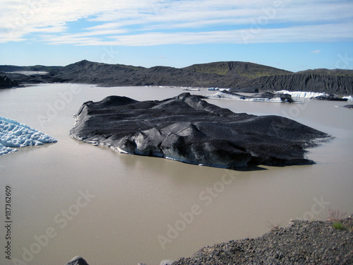 Blick auf einen mit Vulkanasche überzogenen Eishügel in einem Gletschersee in Island