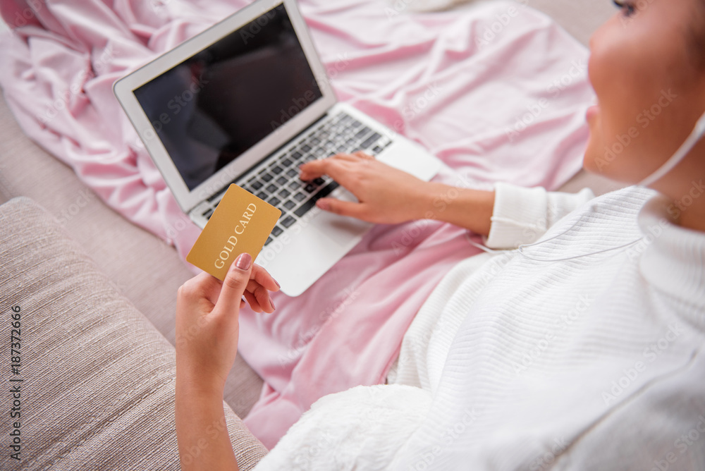 Top view close up of cheerful young woman holding gold credit card in hand. She is sitting on couch and using laptop. Online shopping concept