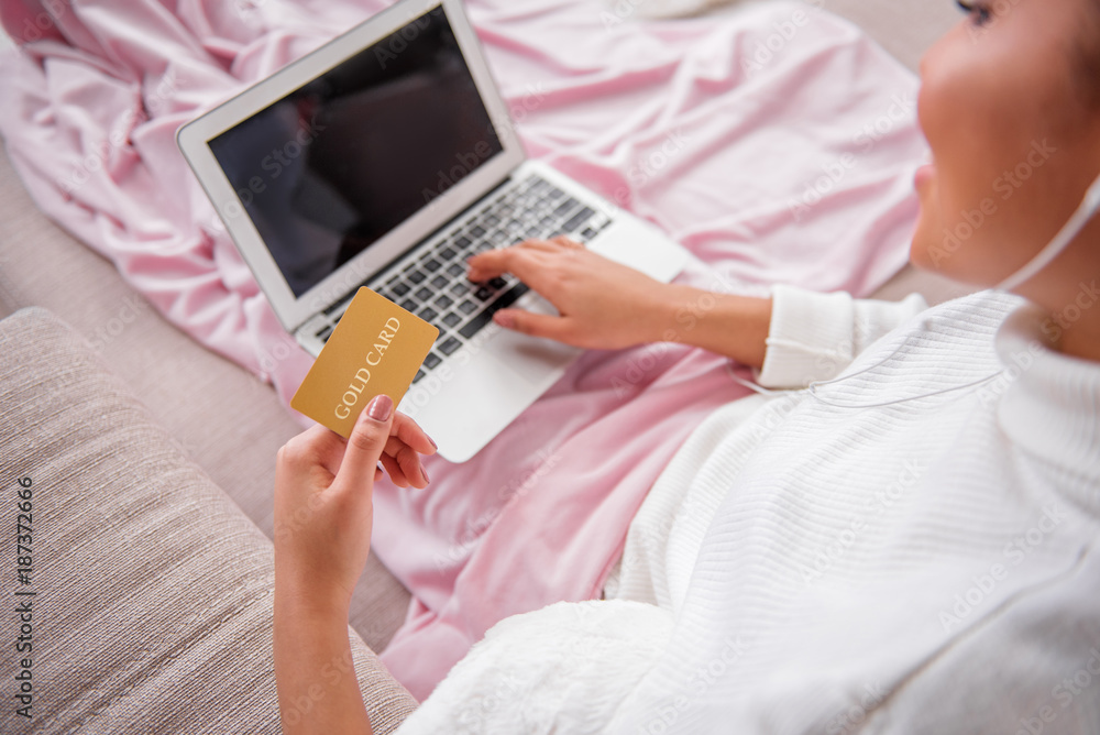 Fototapeta premium Top view close up of cheerful young woman holding gold credit card in hand. She is sitting on couch and using laptop. Online shopping concept