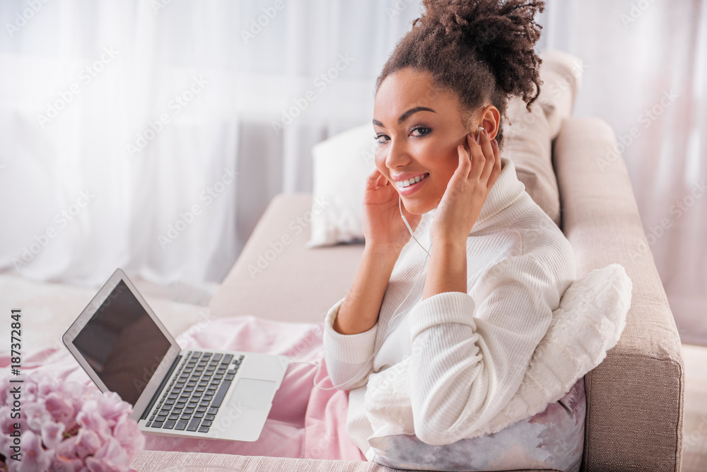 I like music. Portrait of cheerful young woman is touching headphones and smiling. She is looking at camera with excitement while keeping laptop on knees I like music. Portrait of cheerful young woman is touching headphones and smiling. She is looking at camera with excitement while keeping laptop on knees