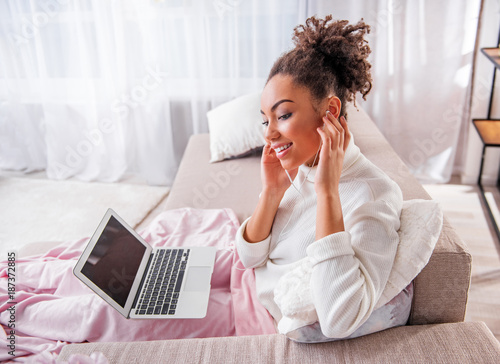Carefree african girl enjoying favorite song on laptop. She is sitting in living room and smiling while touching earphones