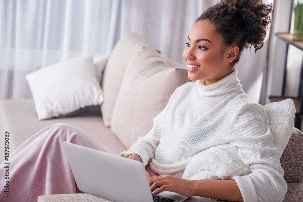 Joyful african girl is working on laptop at home. She is looking aside pensively and smiling. Freelance concept Joyful african girl is working on laptop at home. She is looking aside pensively and smiling. Freelance concept