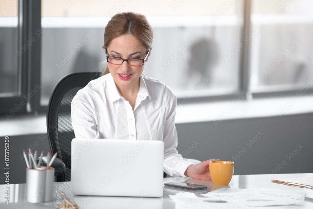 Portrait of smiling lady looking at laptop while tasting mug of tea at desk in office. Labor concept Portrait of smiling lady looking at laptop while tasting mug of tea at desk in office. Labor concept