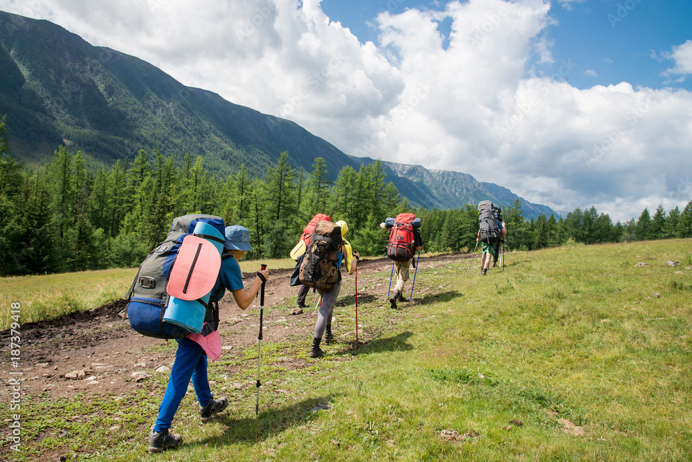 Group of travelers with backpacks walk along a trail towards a mountain ridge by sunny day. Backpackers and hikers style, active leisure