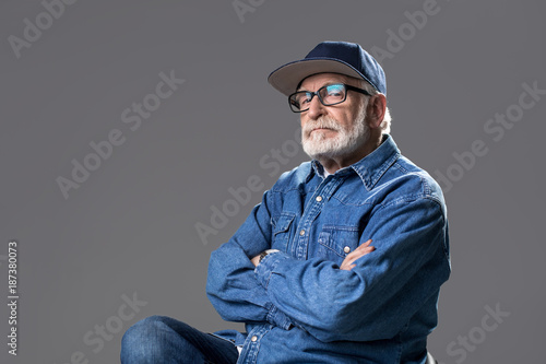 Want to look younger. Waist up portrait of bearded gaffer sitting on chair and looking at camera with pride. He is wearing a cap and glasses. Isolated on grey background