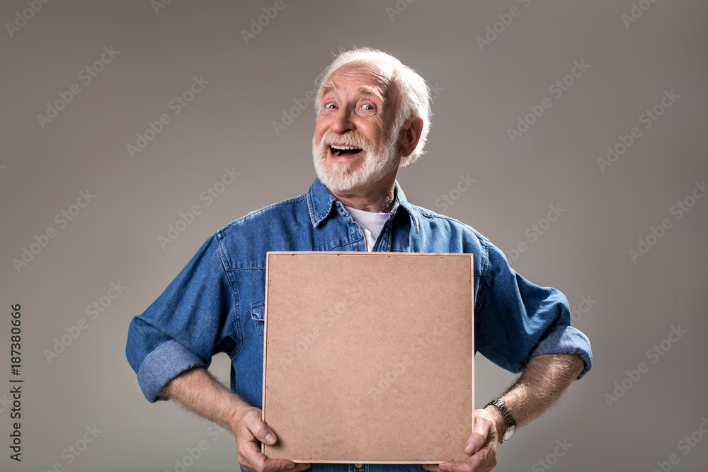 Waist up portrait of humorous elderly man holding wooden frame. Isolated on grey background
