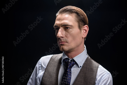 Formidable look. Serious young solid businessman is standing and looking aside confidently. Isolated on dark background