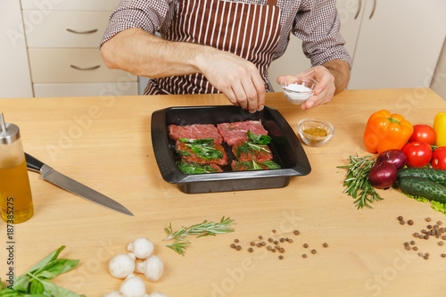Close up young man in apron sitting at table with vegetables, cooking at home preparing meat stake from pork, beef or lamb, salt meat herbs in black baking tray, in light kitchen with wooden surface.