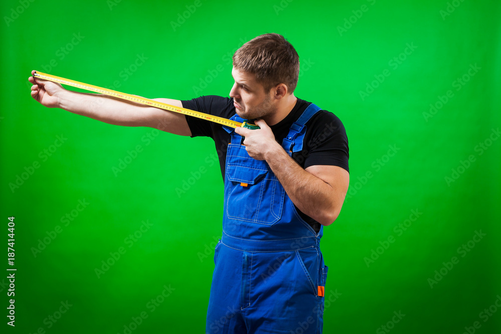 A dark-haired male builder in a black T-shirt and blue construction jumpsuit demonstrates and measures with his long yellow tape rule his hand on a green isolated background A dark-haired male builder in a black T-shirt and blue construction jumpsuit demonstrates and measures with his long yellow tape rule his hand on a green isolated background