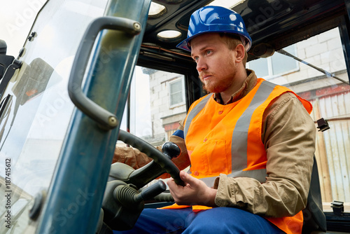 Confident bearded worker wearing protective helmet and reflective vest driving lift truck while working in port warehouse, portrait shot