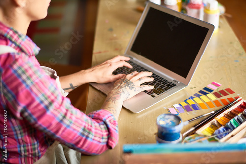 Close up view of creative young woman using modern laptop, focus on tattooed female hands typing and artists supplies on wooden table