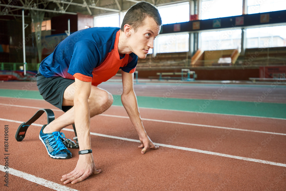 Motivational side view portrait of young amputee athlete on start position on running track in modern indoor stadium, copy space