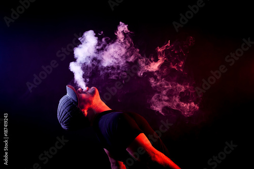 A young man exhales a cloud of colored smoke of red color on a black isolated color all the back is covered with yellow smoke. Harm to smoking on the example of a young mugger