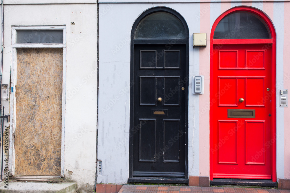 Black door next to a red door and a wooden panel replacing a missing door in the front facade of a Victorian British English house