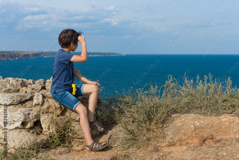 The boy sits on the high seashore and looks out into the distance.