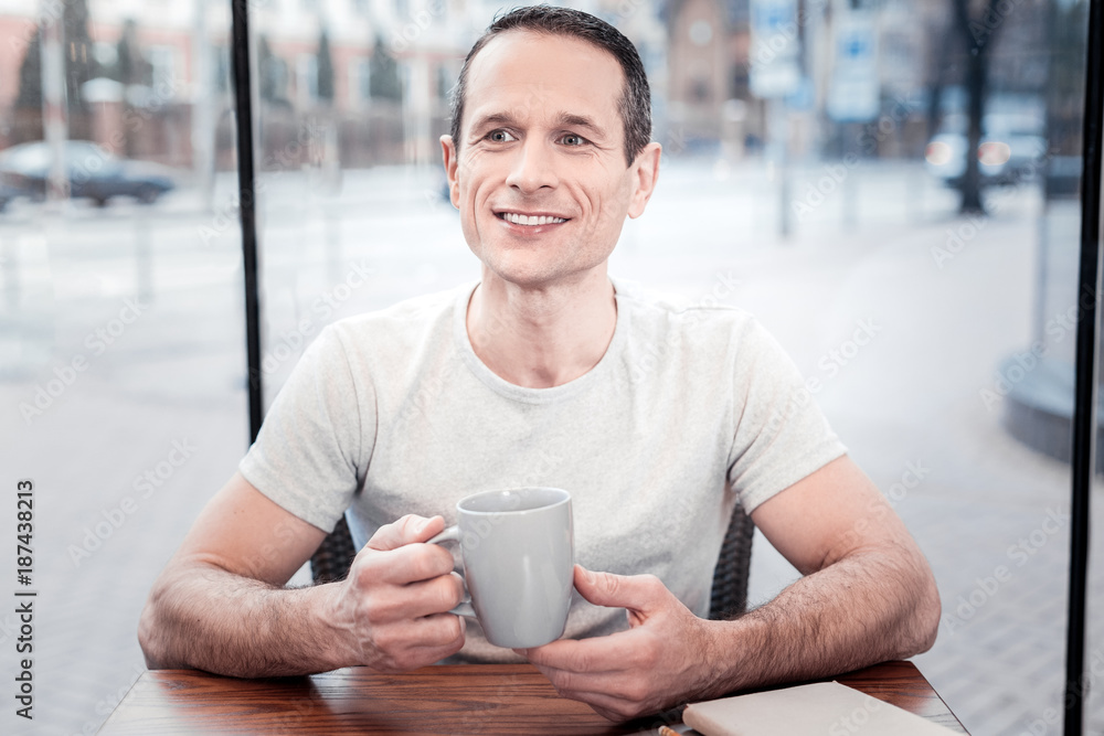 Positive thoughts. Handsome man keeping smile on his face and holding cup in right hand while going to drink tea Positive thoughts. Handsome man keeping smile on his face and holding cup in right hand while going to drink tea